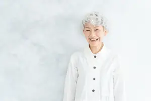 An older woman with white hair and a white shirt with buttons is smiling in front of a white wall.