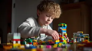 A young boy with curly blonde hair is building a Lego tower on a table.