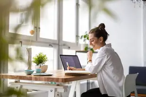 A woman sitting at a table with a laptop and a notebook smiling and holding a pen