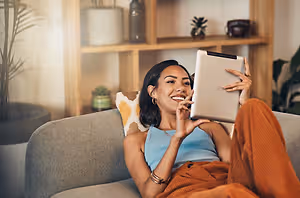 A woman is smiling and holding a tablet in her hands while sitting on a couch and seems to be posing for a photo.