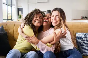 Three women sitting on a couch and hugging each other, smiling and laughing