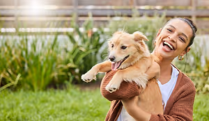A woman holding a dog in a yard with green grass and plants in the background