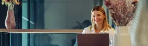 A woman is sitting on a chair in front of a desk with a laptop and smiling at the camera