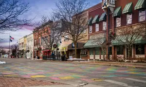 A colorful chalk drawing is on the road with trees and buildings on the side of the road, along with the flag on a pole and street lights. The buildings have awnings, and some have Christmas decorations. A trash bin is in front of a building with a clock on top of the pole. A tree with branches without leaves is on the side of the road. The sky is blue with clouds and a pink hue at the end.