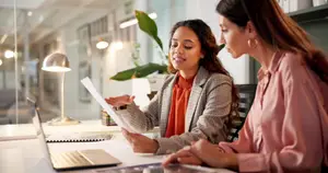 Two women are sitting at a desk in an office with a laptop, papers, and a lamp on the desk.