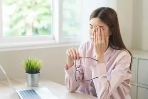 A woman sitting at her desk rubbing her eyes and holding her glasses