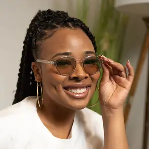 A smiling woman is adjusting her eyeglasses, with a plant and a lamp in the background.