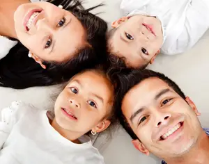 A happy family of four lies on a bed with white sheets, all smiling and looking at the camera. The woman on the left has long black hair and earrings. The man on the right has a smile on his face. The girl in the middle has a mole on her cheek, and the boy on the right has a mole on his cheek. The boy on the left has a mole on his cheek.