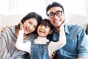 A family of three, consisting of a man, a woman, and a child, are smiling and posing for a picture in a living room with a couch behind them.