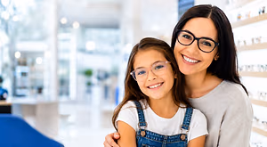 A woman and a young girl are standing next to each other in an optician's shop, both wearing glasses and smiling.