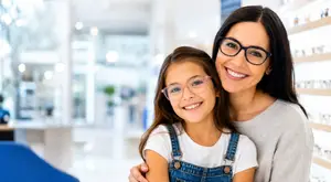 A woman and a young girl are standing next to each other in an optician's shop, both wearing glasses and smiling.