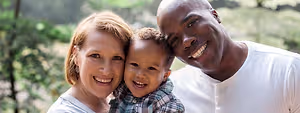 A family of three, consisting of a mother, father, and young boy, are smiling and posing for a photo in a lush green forest background. The boy is holding onto his father's shoulder while the mother stands beside them, all of them dressed in white shirts and looking joyful and happy.