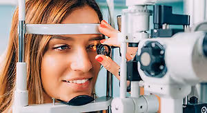 A woman is looking into an ophthalmoscope machine, probably in an examination room.