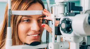 A woman is looking into an ophthalmoscope machine, probably in an examination room.