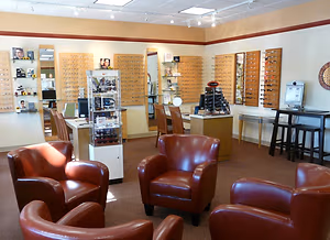 A modern waiting room with leather chairs, wooden tables, mirrors, and shelves with eyewear. The room features ceiling lights and a desk with a computer monitor.