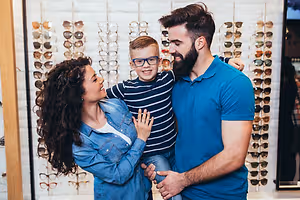 A young boy is sitting on his fathers lap while looking at sunglasses with his mother in an optical shop