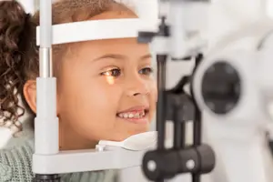 A young girl wearing a green sweater is looking through a white and black ophthalmoscope.