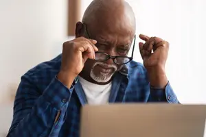 African American man in blue shirt adjusting his glasses while using a laptop.