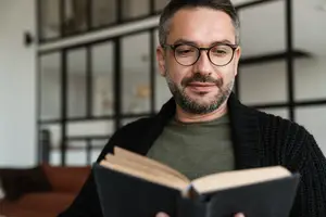 A man is reading a book while wearing glasses and a black cardigan