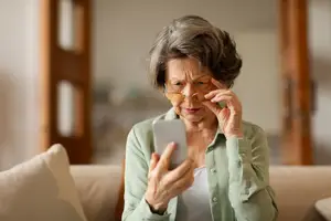 An elderly woman wearing eyeglasses and a light green shirt is sitting on a couch and using her phone.