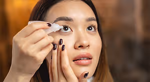 A woman is applying eye drops to her eyes while wearing a white shirt and has brown hair.