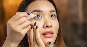 A woman is applying eye drops to her eyes while wearing a white shirt and has brown hair.