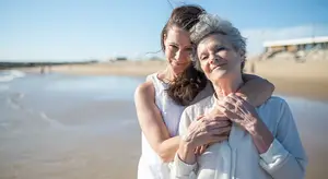 Two women standing on a beach, one holding the other's shoulder, smiling.
