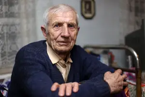 An elderly man with white hair wearing a blue sweater and sitting on a bed in a room with a white wall and a picture frame