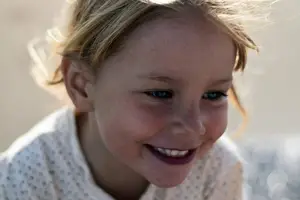 A young girl with blonde hair is smiling and looking at the camera with a slightly open mouth