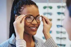 A woman with glasses smiles at another person in a shop with glasses on display behind her
