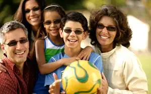 A family is standing close to each other and smiling in front of a white building. They are wearing sunglasses, and the man is holding a soccer ball. The woman is wearing a necklace, and the little girl is holding a soccer ball. The background is blurry, and there are trees and plants.