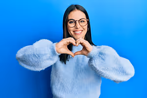 A woman wearing a blue sweater and glasses forms a heart shape with her hands and smiles for a photo in front of a blue background.