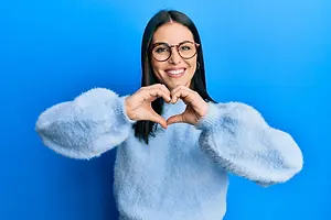 A woman wearing a blue sweater and glasses forms a heart shape with her hands and smiles for a photo in front of a blue background.