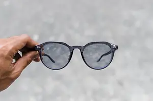 A hand holds a pair of glasses with black frames and lenses, showing the details of the glasses and the blurred background behind the hand.