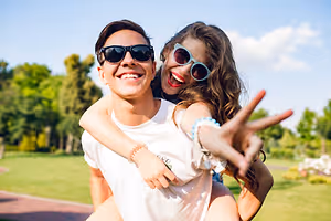 A man and a woman are smiling while posing for a photo in a park, the woman is making a peace sign with her hand
