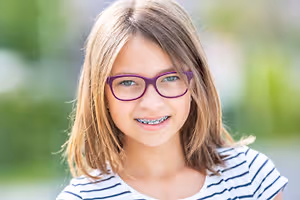 A young girl with braces and glasses is smiling and looking forward, possibly posing for a photo.
