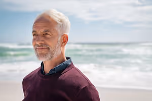 A smiling man in a maroon sweater standing on a beach with the ocean in the background