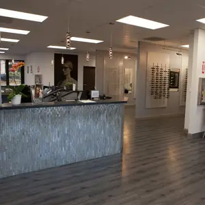 A photo of an empty room with a counter, potted plant, photo frame, and large poster of a woman wearing glasses is on the wall. The room has white walls, a tiled floor, and a ceiling with lights and hanging lamps. There are also eyeglasses displayed on the wall and a white column with a photo frame on top. There is a glass door with a glass window and a potted plant on the left side.