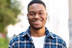 A young black man wearing a blue and white checkered shirt and white earphones with a smile on his face, standing on the street and posing for a photo