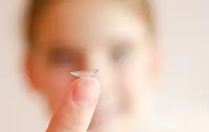 A woman holds a contact lens in her hand, with a blurred background and another person's face in the distance.