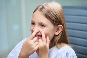 A young girl with blonde hair sits in a chair with her hands on her nose, wearing a white shirt and looking at something in front of her.