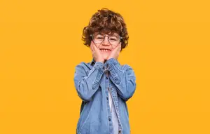 Happy young boy with curly hair wearing glasses and a blue denim jacket posing for a photo against a yellow background