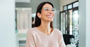 A smiling woman wearing glasses stands in a room with a bright light behind her. She is wearing a light pink sweater and has a warm expression on her face. The background is a bit blurry, with a white wall and a glass door visible.
