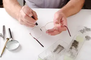 A man wearing a blue plaid shirt is adjusting a pair of glasses on a white table with various tools.