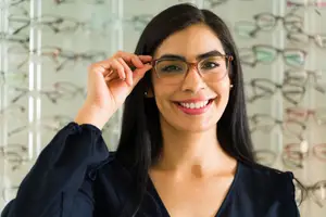 A woman in a dark blue blouse is smiling and holding up a pair of brown-framed glasses in front of her face, with a blurry background of various glasses frames in different colors and styles displayed on a white wall.