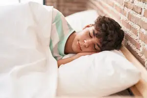 Teenage boy sleeping in a white bed in a room with a brick wall and wooden bed frame.