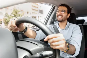 Man wearing glasses and bracelets driving a car with buildings and trees in the background.