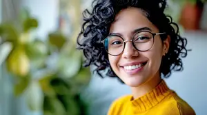 A woman with curly black hair is smiling while wearing glasses and a yellow turtleneck sweater in front of a blurry green plant.