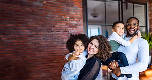 A family of four is posing for a photo in front of a brick wall. The man is wearing eyeglasses and a white shirt, and he is holding a young boy on his shoulder. The woman is wearing a black blouse and is holding a young girl in her arms. The young boy is wearing a green shirt, and the young girl is wearing a white dress. Behind them is a brick wall with a glass window. There is also a potted plant on the floor.