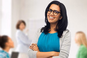Confident woman wearing glasses and a teal top standing in a room with blurred colleagues behind her.